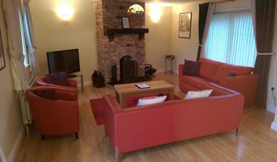 Image shows lounge area with sofas, chairs, TV and fireplace. Two windows in the room.