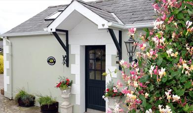 Images shows front door of cottage with hedge and flowers to the left of it.