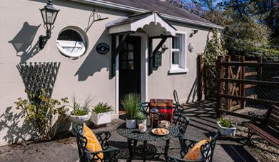 Image is of patio area at back of cottage with wrought iron table and 2 chairs.