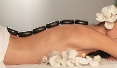 Image is of a lady with stones on her back for a hot stone massage. A display of white orchids are also in the image
