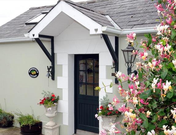 Images shows front door of cottage with hedge and flowers to the left of it.
