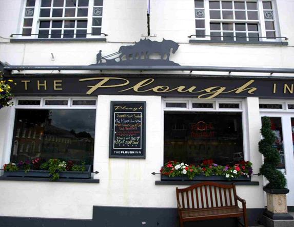 Image is of the outside of the restaurant with a wooden bench outside one of the windows and bay trees at either side of the front door