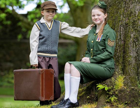 Image shows girl in military uniform sitting under a tree with a young boy wearing a cap standing beside her holding a brown suitcase