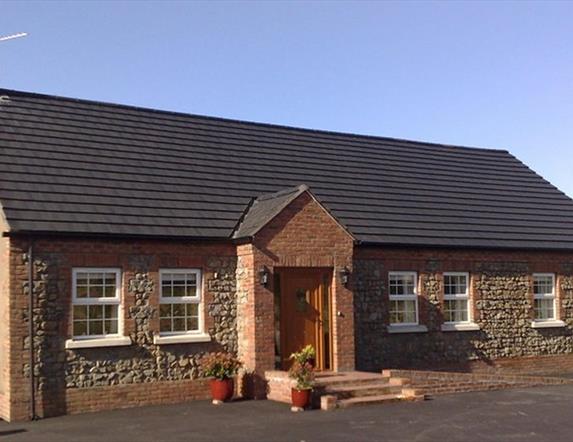 Image shows front of detached cottage which has 6 windows and front door with a few low steps