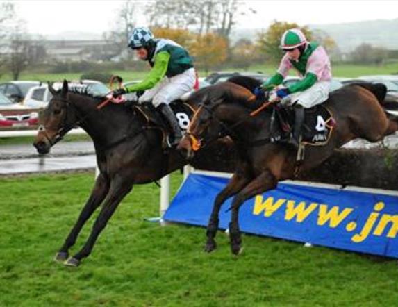 Image shows two jockeys on horses jumping over the fence