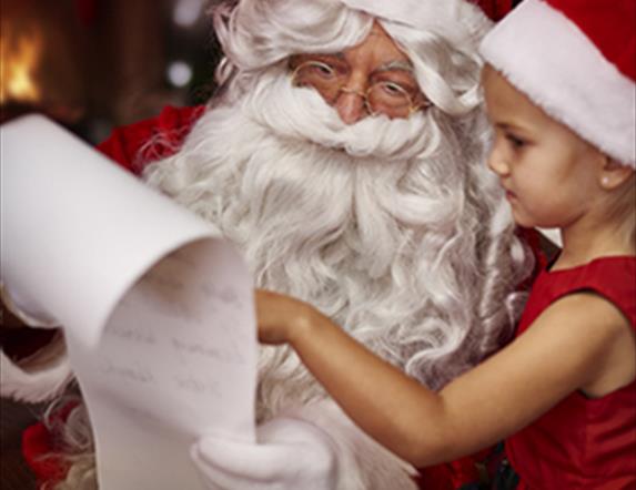 image shows picture of santa claus and a little girl in a red and white santa hat and red dress, looking at santas long list on a scroll of white pape