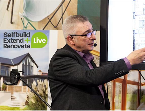 Image is of exhibitor pointing at a set of house plans at the Selfbuild Extend Renovate exhibition in Eikon Exhibition Centre Lisburn