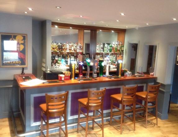 Image shows bar area with 4 bar stools on wooden bar bench and bar is painted a soft lilac colour with recessed lighting overhead.