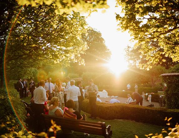 Image shows a beautiful garden with people gathering around and the sun shining through the trees