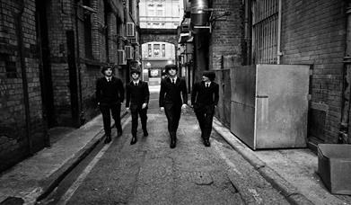 Black and White Image shows Beatles Tribute members in a alleyway dressed in suits.