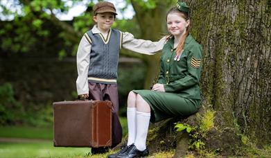 Image shows girl in military uniform sitting under a tree with a young boy wearing a cap standing beside her holding a brown suitcase