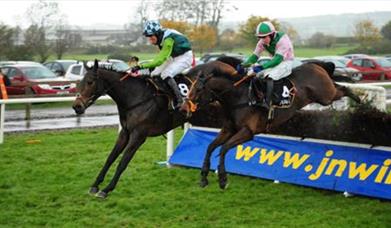 Image shows two jockeys on horses jumping over the fence