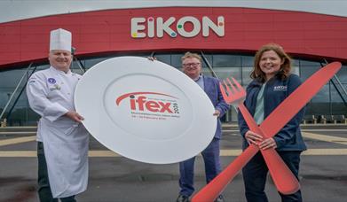 Image shows red Eikon Banner with white text in background with a chef and man holding a large plate and a lady holding large red knife and fork