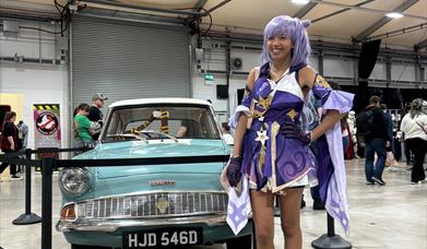 Image is of female dressed up for Comic Con standing beside an old car