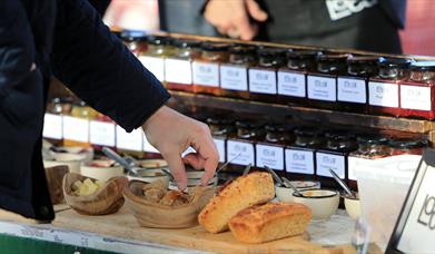 Image shows rows of jars of relish or jam and a customer's hand lifting a piece of bread to taste