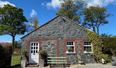 Image shows stone cottage covered in ivy at the right hand side plus white front door and bench outside