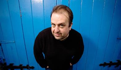 Image is of the comedian Karl Spain smiling and standing in front of a blue wooden door