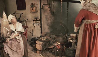 Image is a scene from a weaver's cottage where women are cooking on a fire and one woman is weaving linen