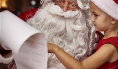image shows picture of santa claus and a little girl in a red and white santa hat and red dress, looking at santas long list on a scroll of white pape