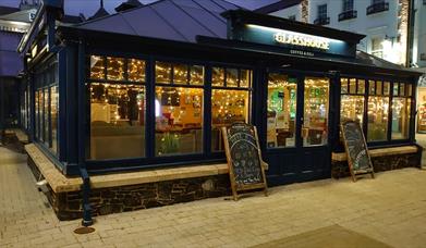 Image shows picture of the front of the glasshouse cafe 
windows with lights inside and two chalkboards leaning against premises.