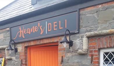 Image shows picture of the front of Keanes Deli stone building and part of the orange front door.  The signage has a grey background with orange colou