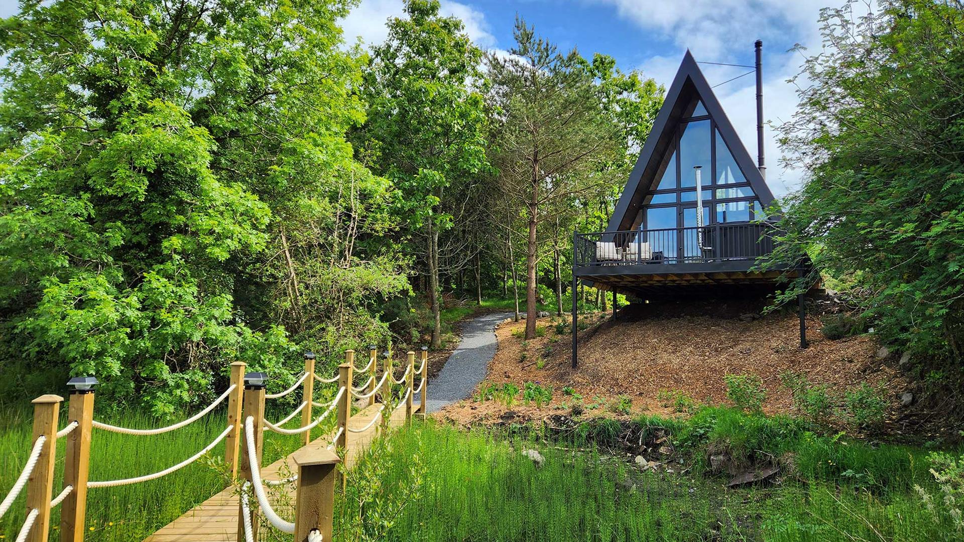 A luxury A-frame lodge set in peaceful woodland surroundings, overlooking a pond with a wooden rope bridge leading through the trees.