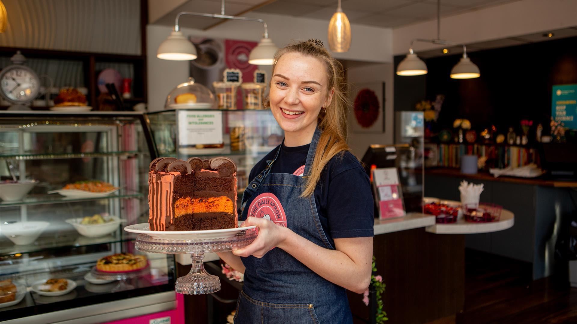 Image shows smiling member of staff holding a large  chocolate and orange cake