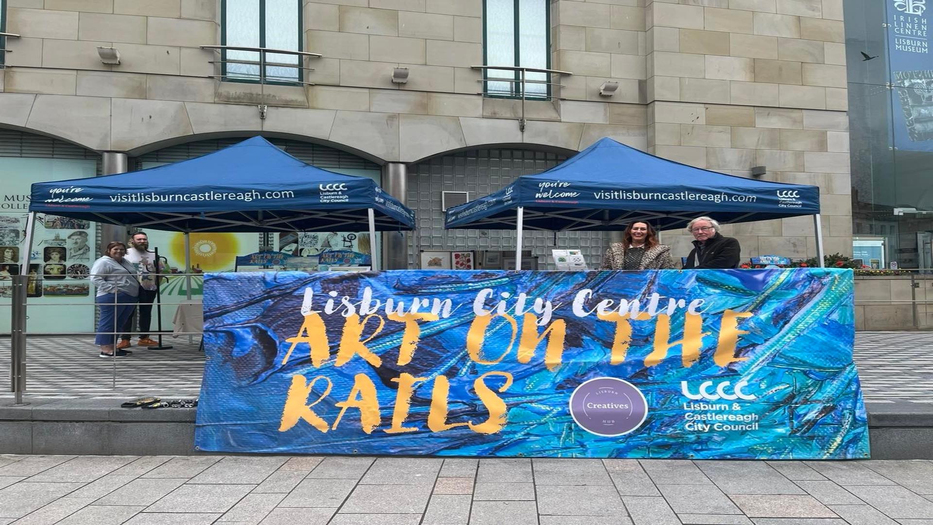 Image shows artists from Art On The Rails standing outside the Irish Linen Museum behind a blue poster, with gazebos and art displayed.