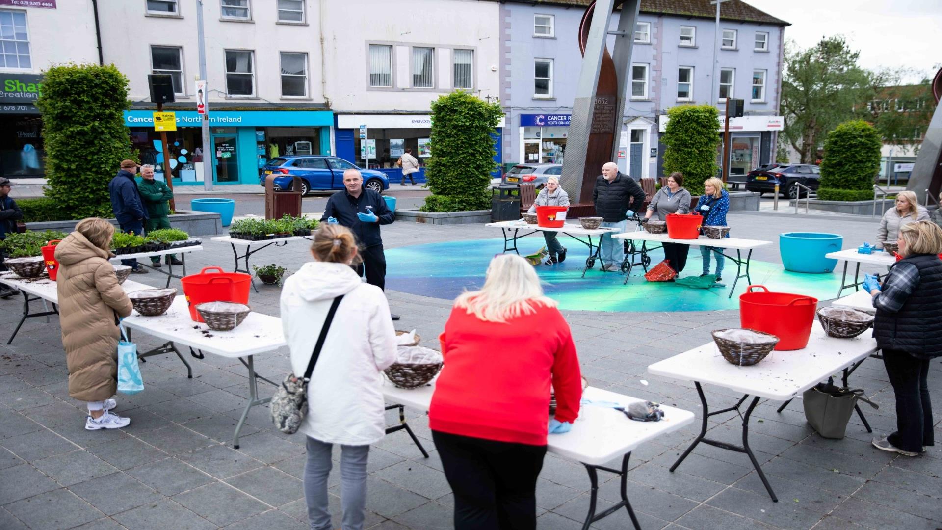 Image shows tables with hanging baskets and members of the public listening to one of the gardeners on how best to design them.