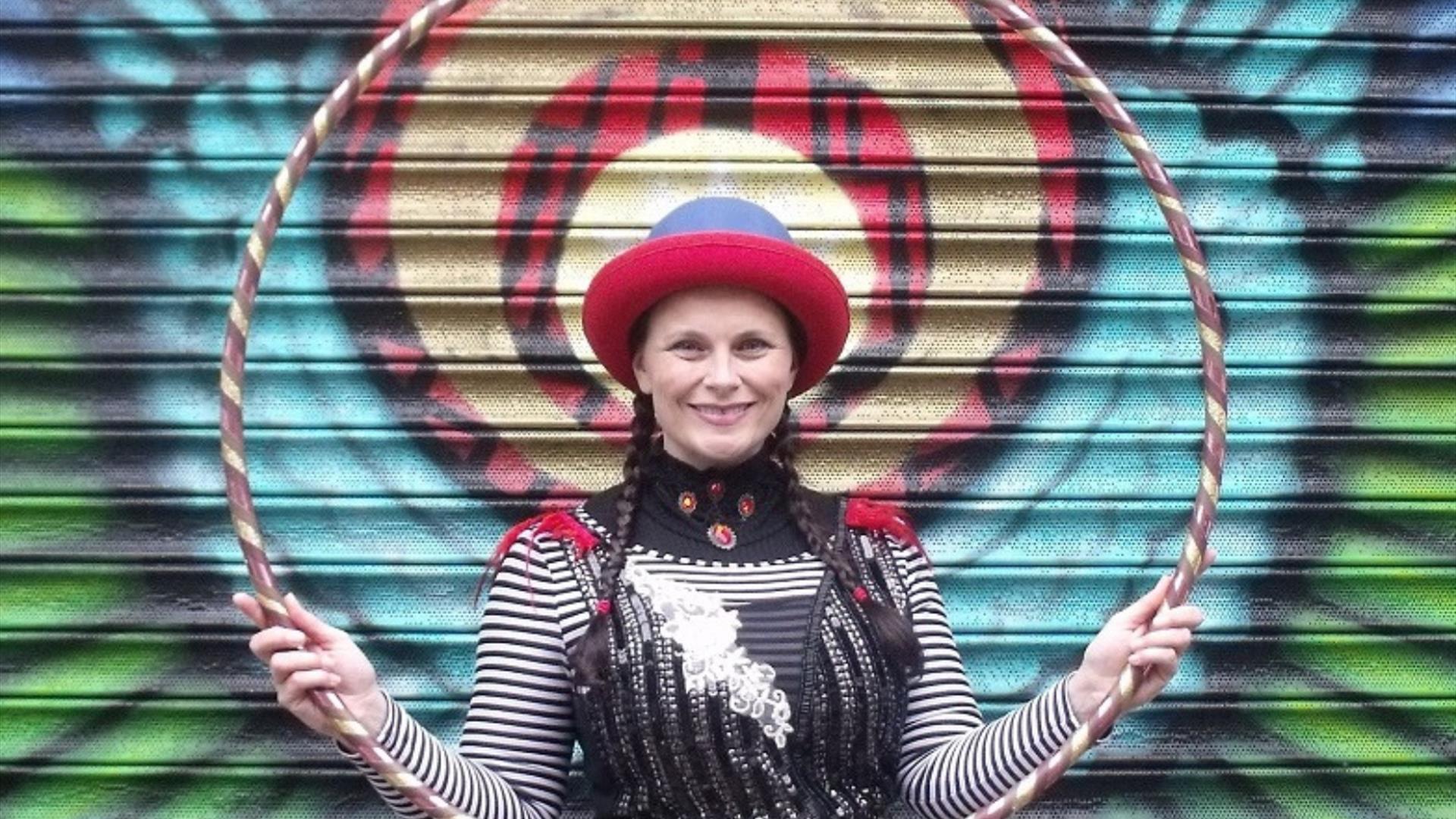 Woman in costume holding a hula hoop in front of a painted door. Circusful performer. Friday Fun in the Park. Moira Demesne.