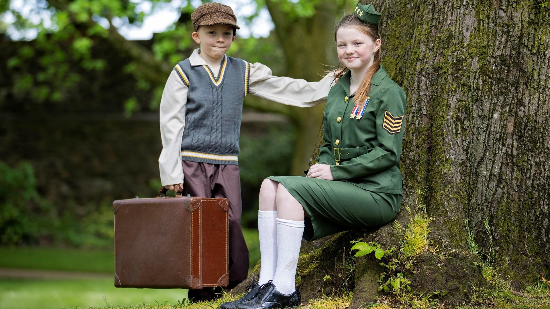 Image shows girl in military uniform sitting under a tree with a young boy wearing a cap standing beside her holding a brown suitcase