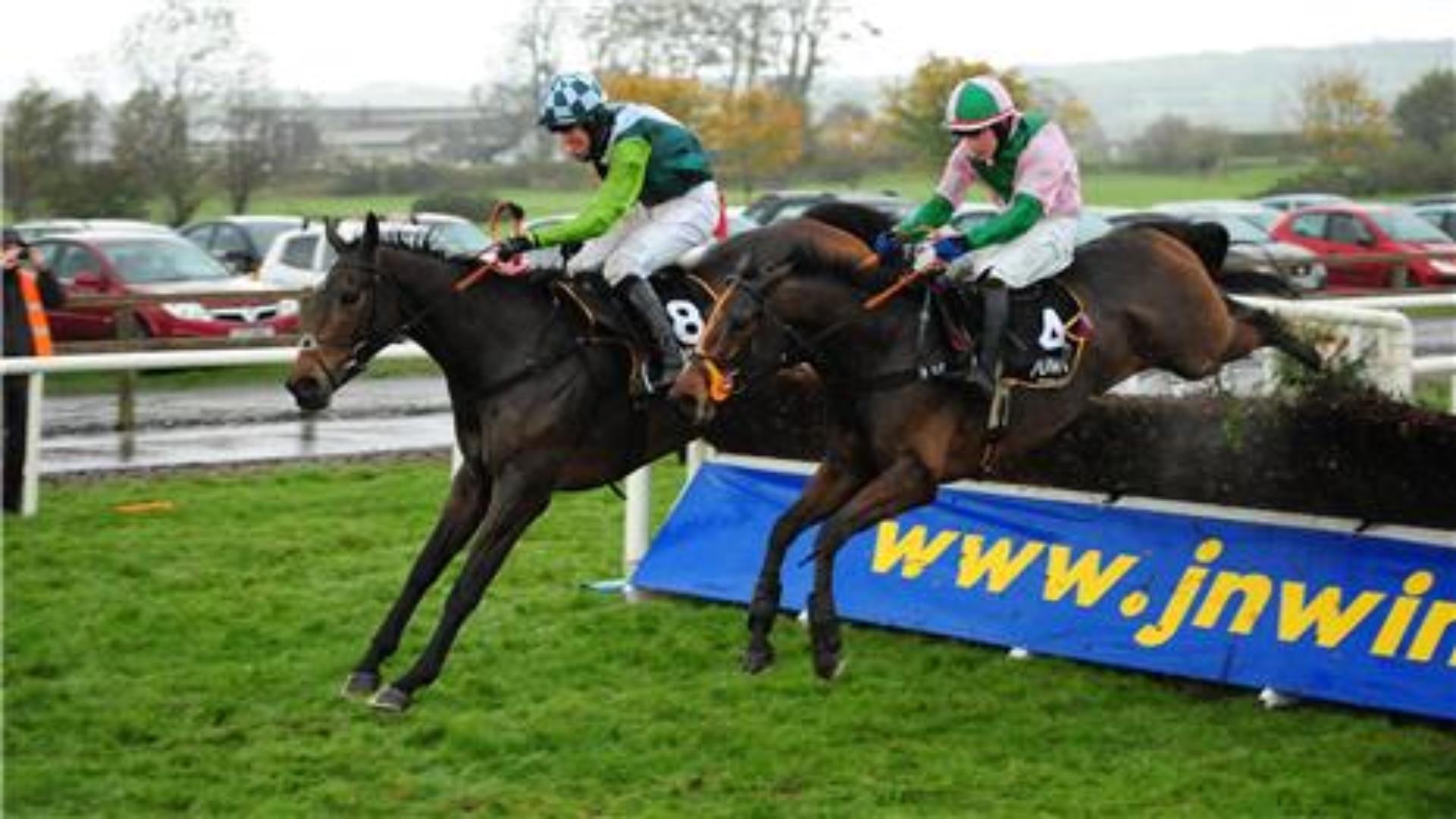 Image shows two jockeys on horses jumping over the fence