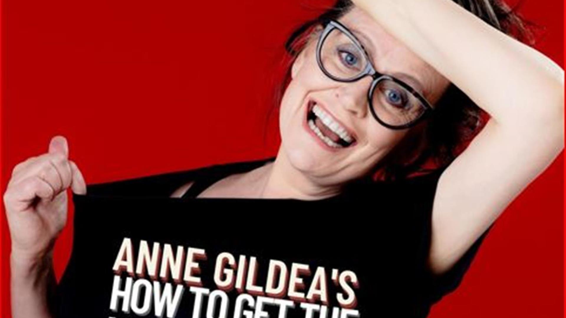 Picture of Anne Gildea holding a microphone above her head with a black top with the saying Anne Gildea's How to get the menopause & enjoy it