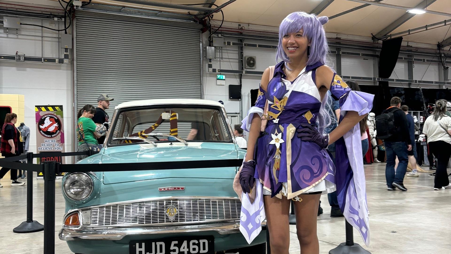 Image is of female dressed up for Comic Con standing beside an old car