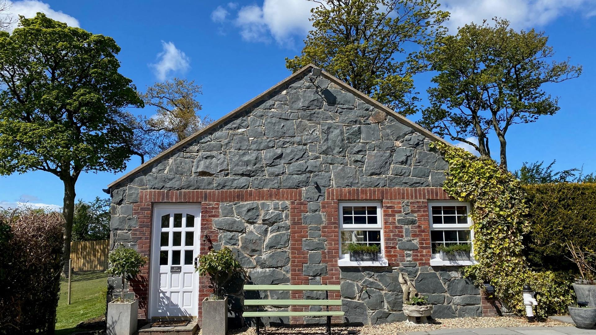 Image shows stone cottage covered in ivy at the right hand side plus white front door and bench outside
