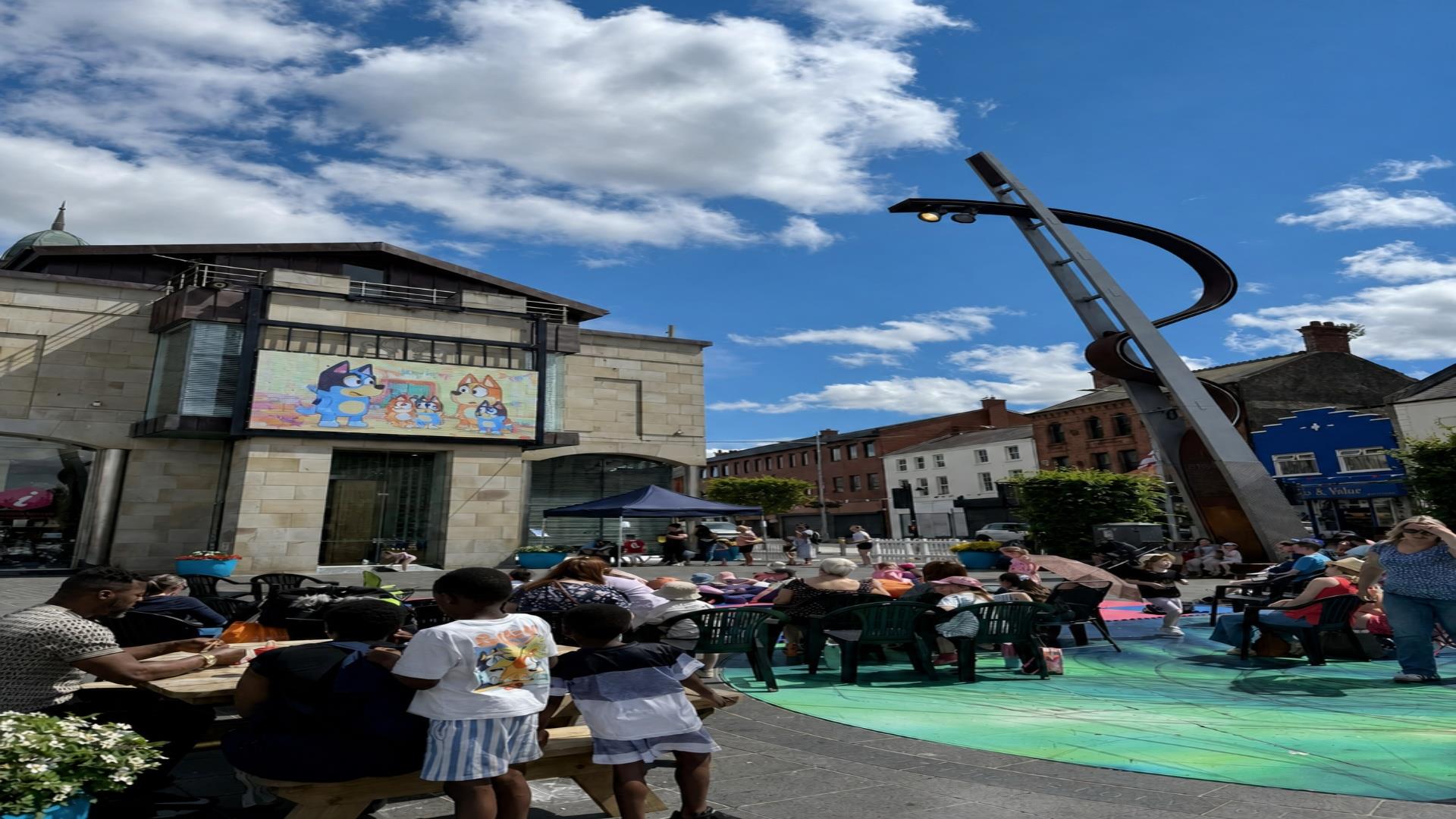 People sitting outside the Irish Linen Museum in Market Square watching a movie on the big screen and enjoying the sunshine.