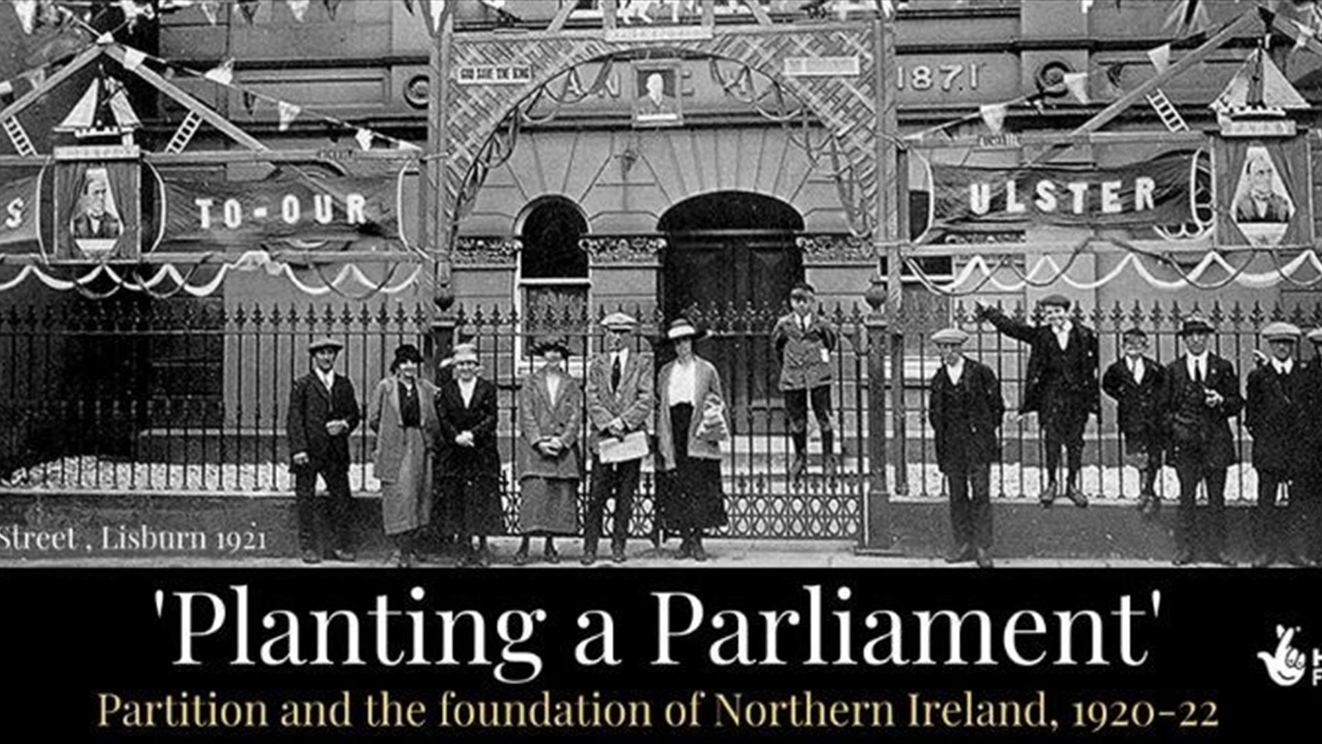 Image shows a black and white photo of group of men, women and children from the 1920s standing outside the gates of a building