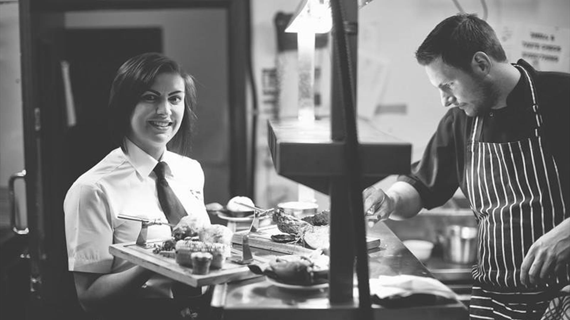 A black and white image showing the chef in the kitchen with a waitress carrying food.