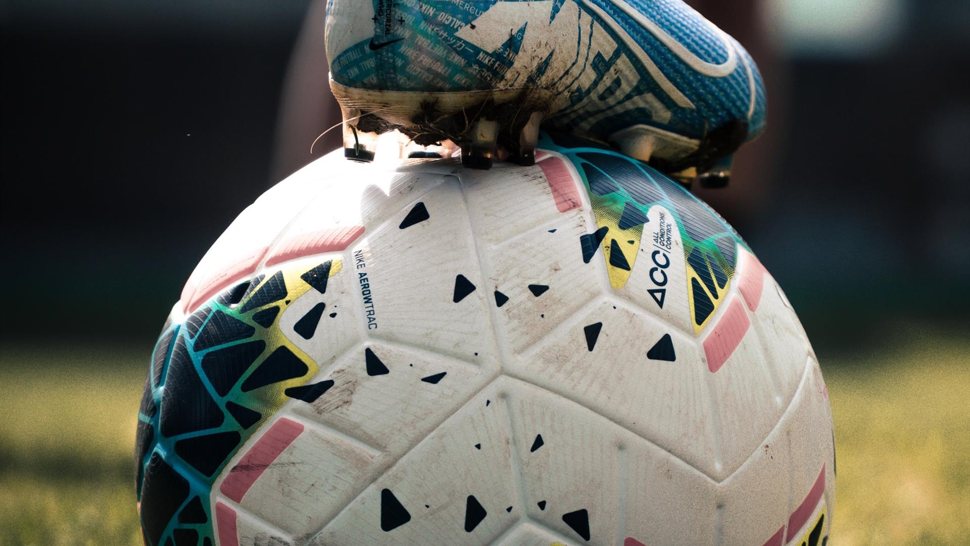 An image of a football with someone holding it in place by their football boot