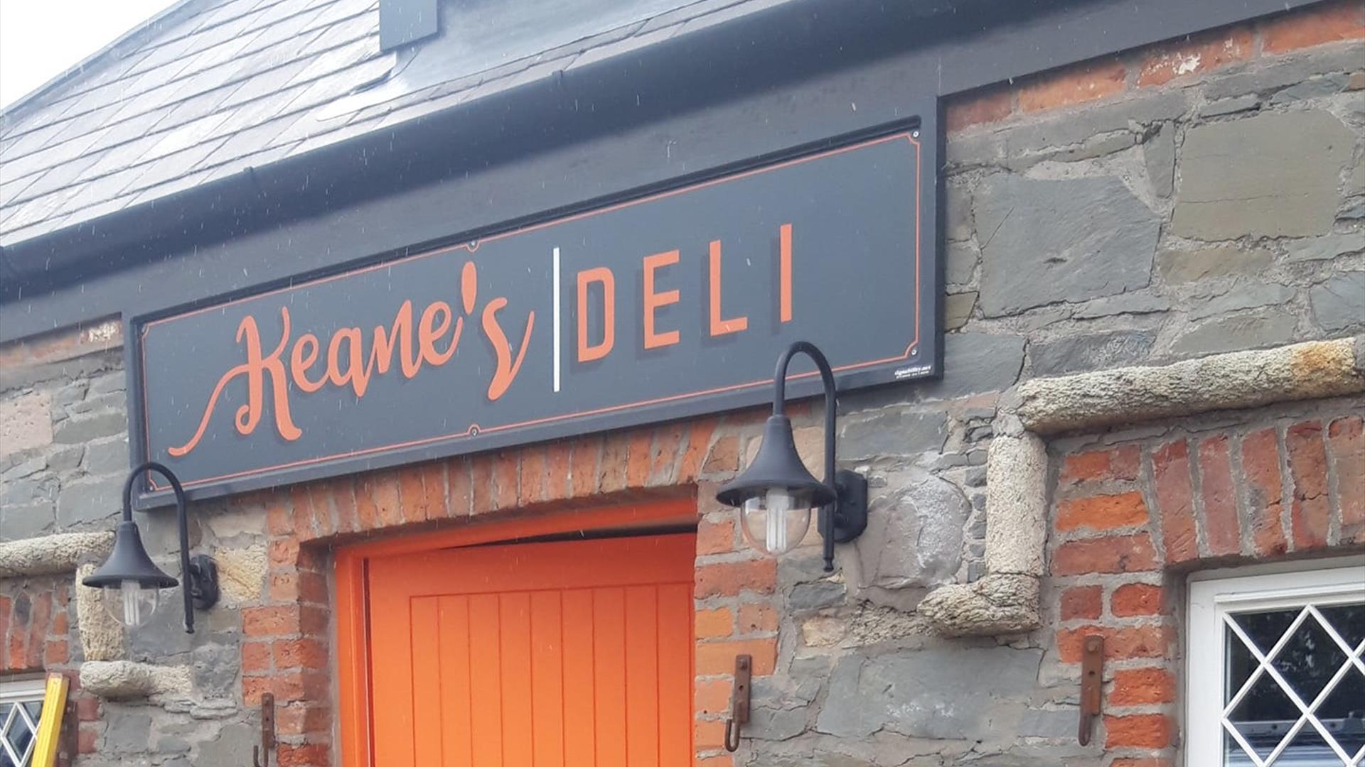 Image shows picture of the front of Keanes Deli stone building and part of the orange front door.  The signage has a grey background with orange colou