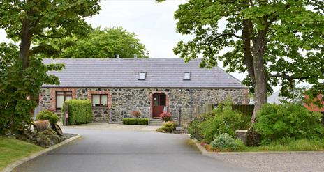 Image shows front of the property. Tarmac driveway bordered by trees on both sides. There is a ramp up to front door and also a path with some low ste