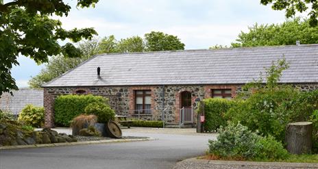 Image shows outside of stone built property with wooden picnic table in small courtyard. Hedges and trees in foreground.