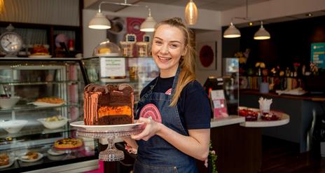 Image shows smiling member of staff holding a large  chocolate and orange cake