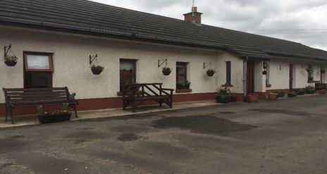 Image shows outside of cottage. Wooden benches outside. Gravel driveway.