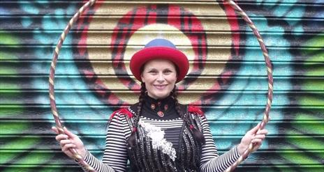 Woman in costume holding a hula hoop in front of a painted door. Circusful performer. Friday Fun in the Park. Moira Demesne.