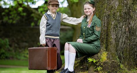 Image shows girl in military uniform sitting under a tree with a young boy wearing a cap standing beside her holding a brown suitcase