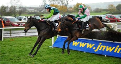 Image shows two jockeys on horses jumping over the fence