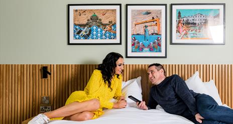 Image shows a man and woman lounging on a double bed with paintings of Northern Ireland on the background wall