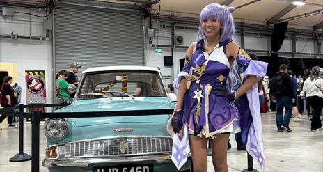 Image is of female dressed up for Comic Con standing beside an old car