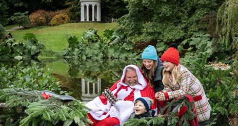 Image shows Santa with 3 children wearing coloured woolly hats laughing & sitting in Hillsborough Castle Gardens.